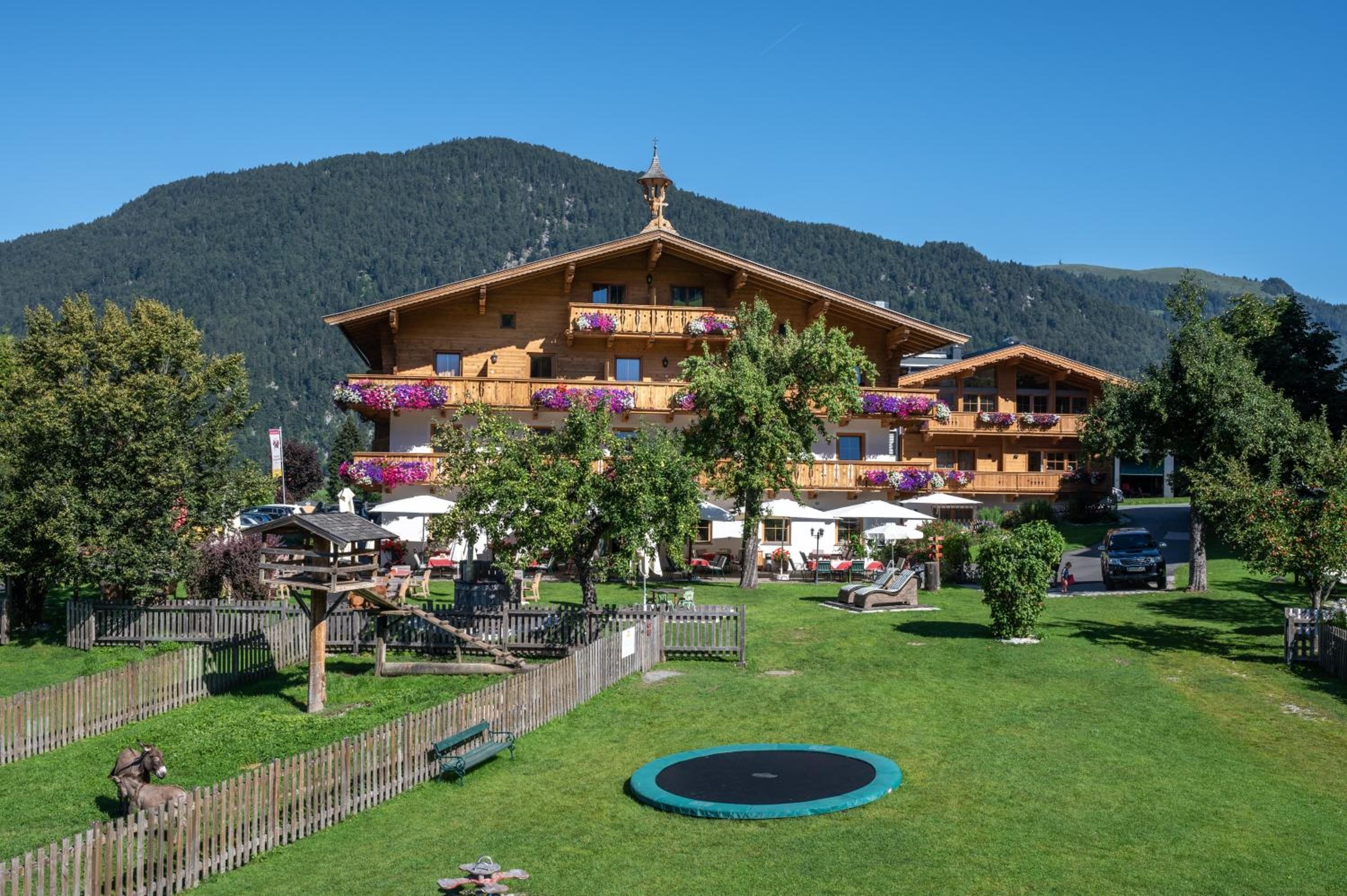 A wooden chalet with balconies, surrounded by a fenced garden, a trampoline, and a mountain backdrop.