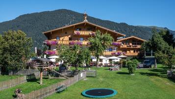 A wooden chalet with balconies, surrounded by a fenced garden, a trampoline, and a mountain backdrop.