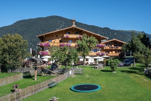 Eine Holzhütte mit Balkonen, umgeben von einem eingezäunten Garten, einem Trampolin und einer Bergkulisse.