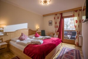A hotel room with a wooden bed, a red blanket, a small table with a lamp, and a view of the outside through a window with curtains.