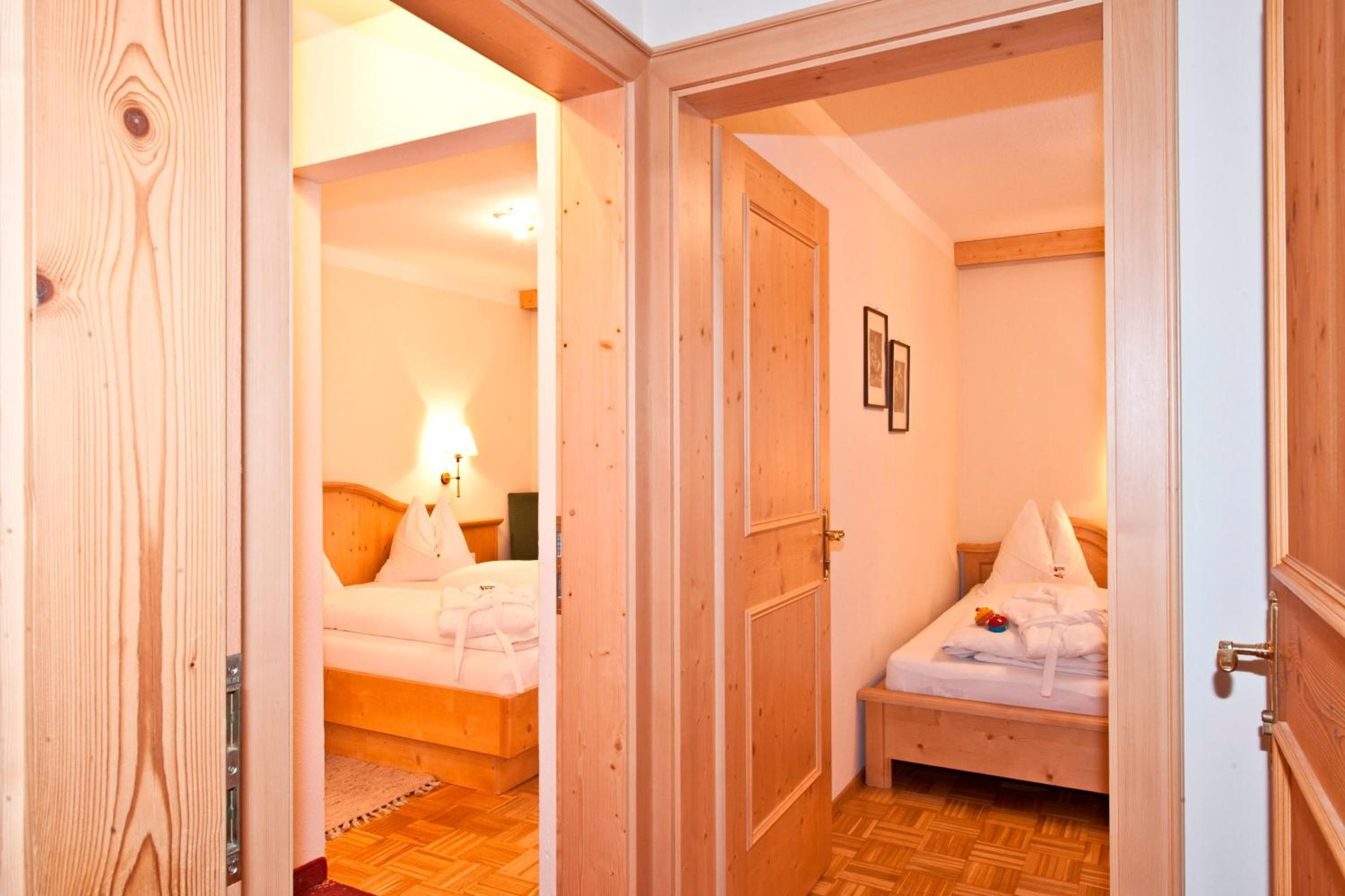 A hotel room with a wooden floor, a bed with white bedding, a bedside table with a lamp, and a door leading to another room.