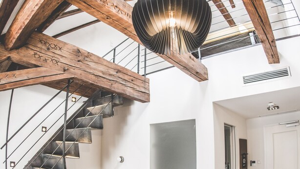 Um amplo loft com vigas de madeira expostas, uma escada moderna, um sofá amarelo e uma luminária pendente única.