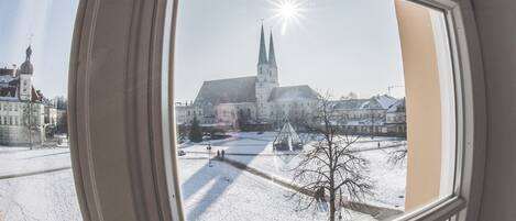 A view through a window of a building with a church in the background, a snow-covered square, and a clear sky.