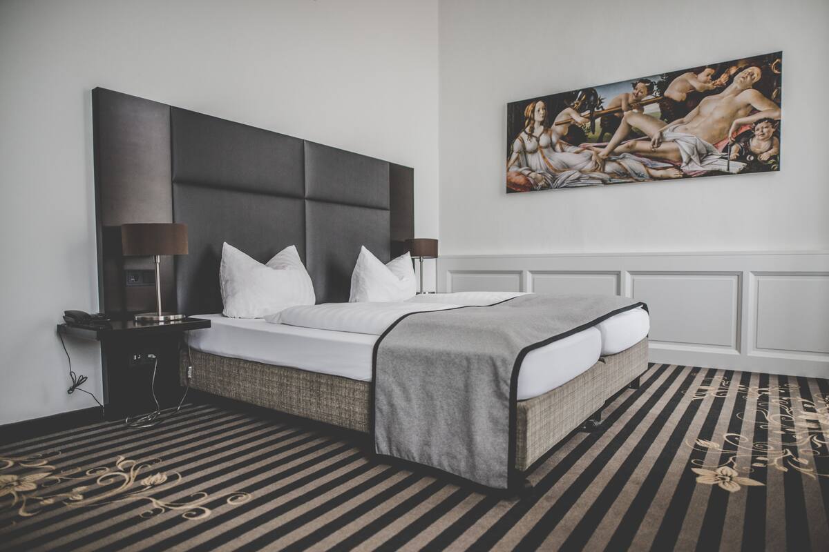A modern hotel room with a large bed, a bedside table, a lamp, a painting on the wall, and a striped carpet.