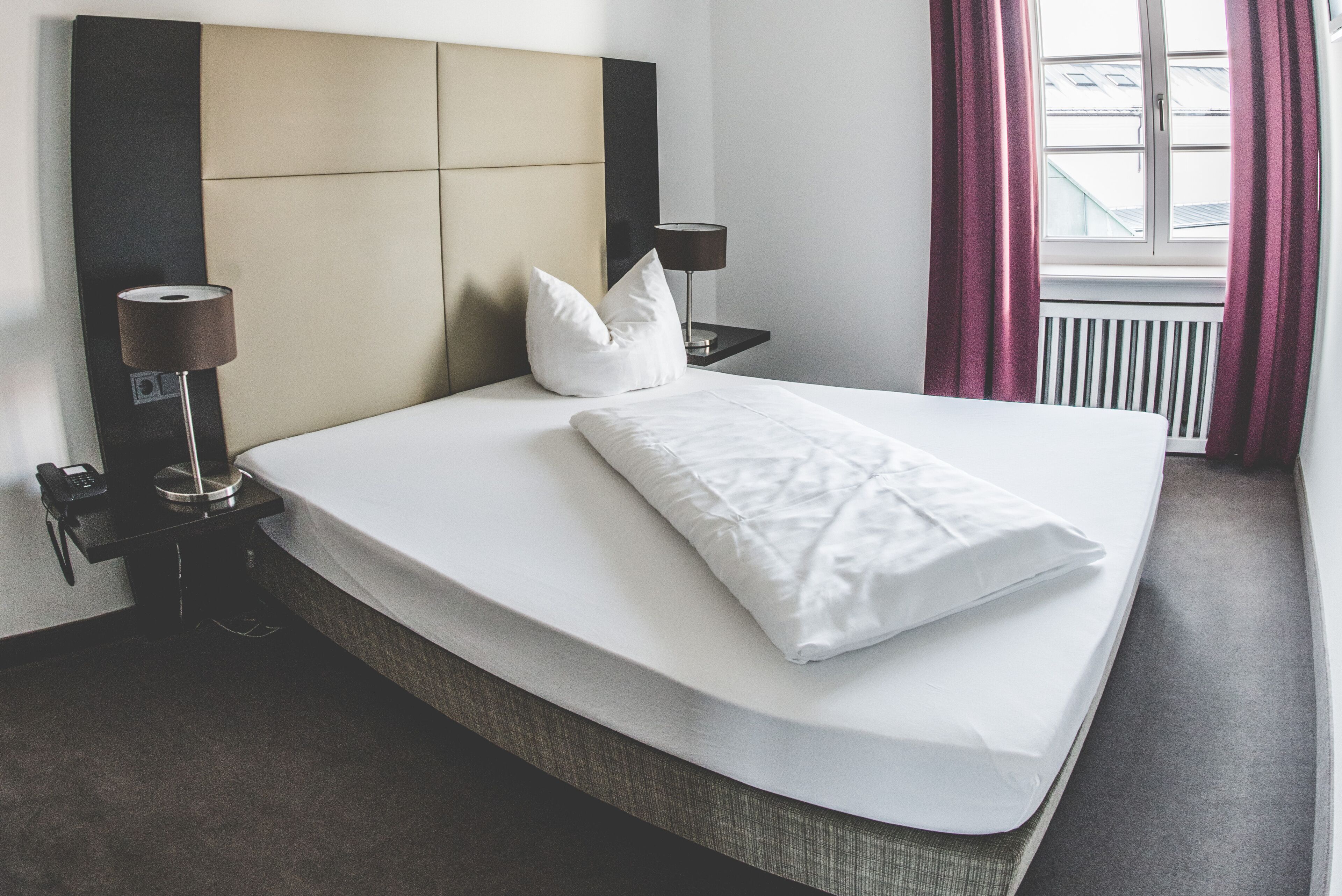 Ein ordentlich bezogenes Bett mit weißen Leinen und Kissen, ein Nachttisch mit Lampe sowie ein Fenster mit Vorhängen.