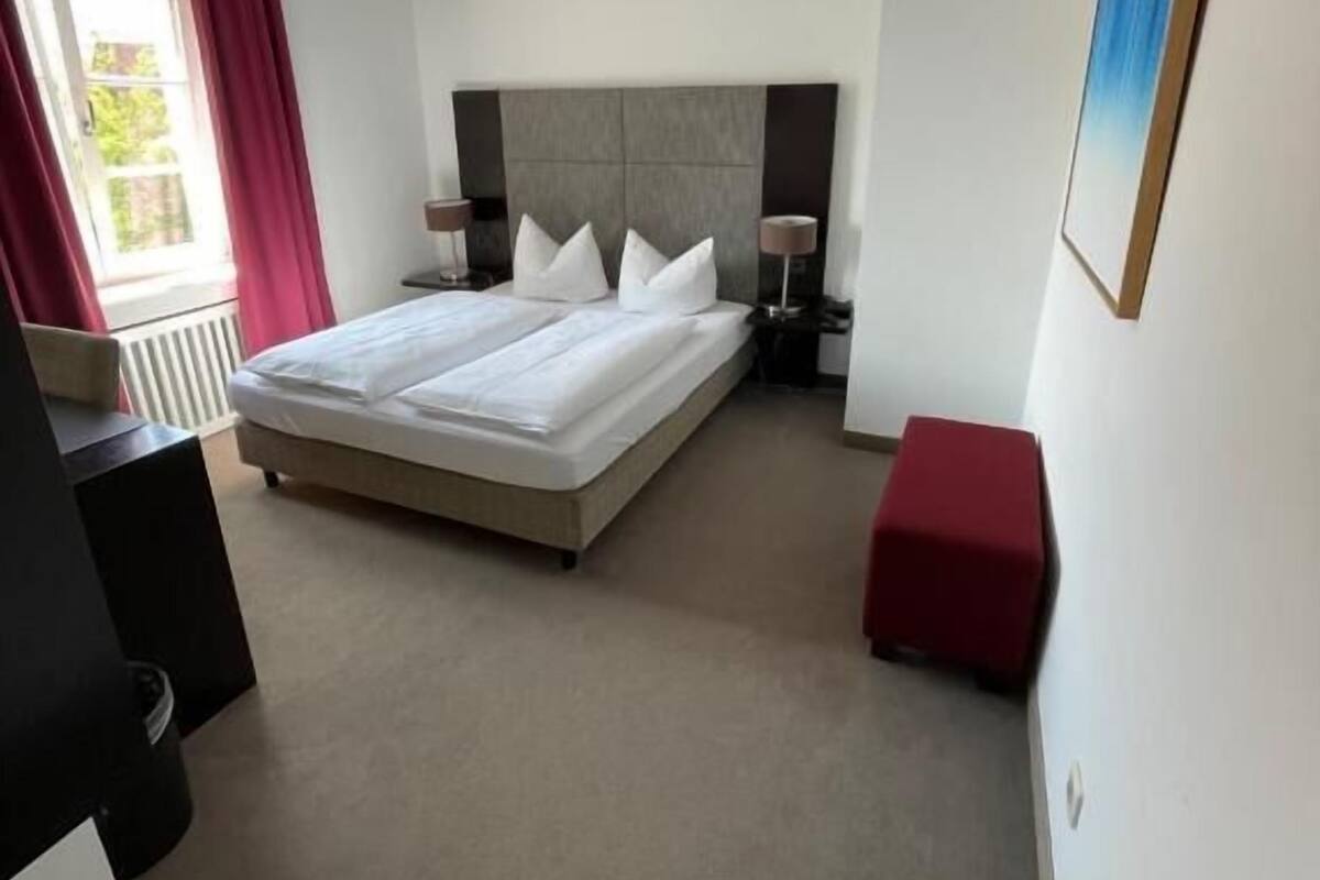 A modern hotel room with a large bed, bedside tables, a red ottoman, and a painting on the wall.
