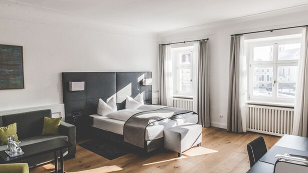 Et moderne hotelværelse med en stor seng, et siddeområde med sofa og bord, samt udsigt over byen gennem store vinduer.