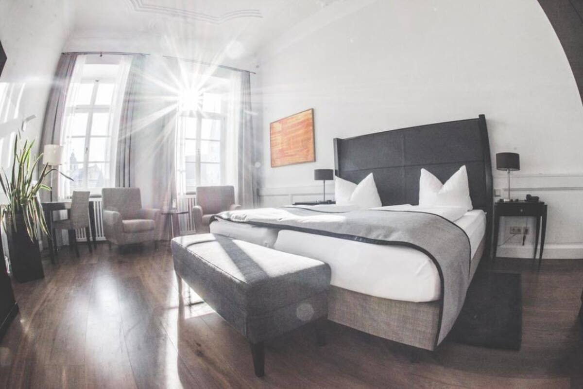A modern hotel room with a large bed, a seating area with chairs, a small table, and a large window with sunlight streaming in.