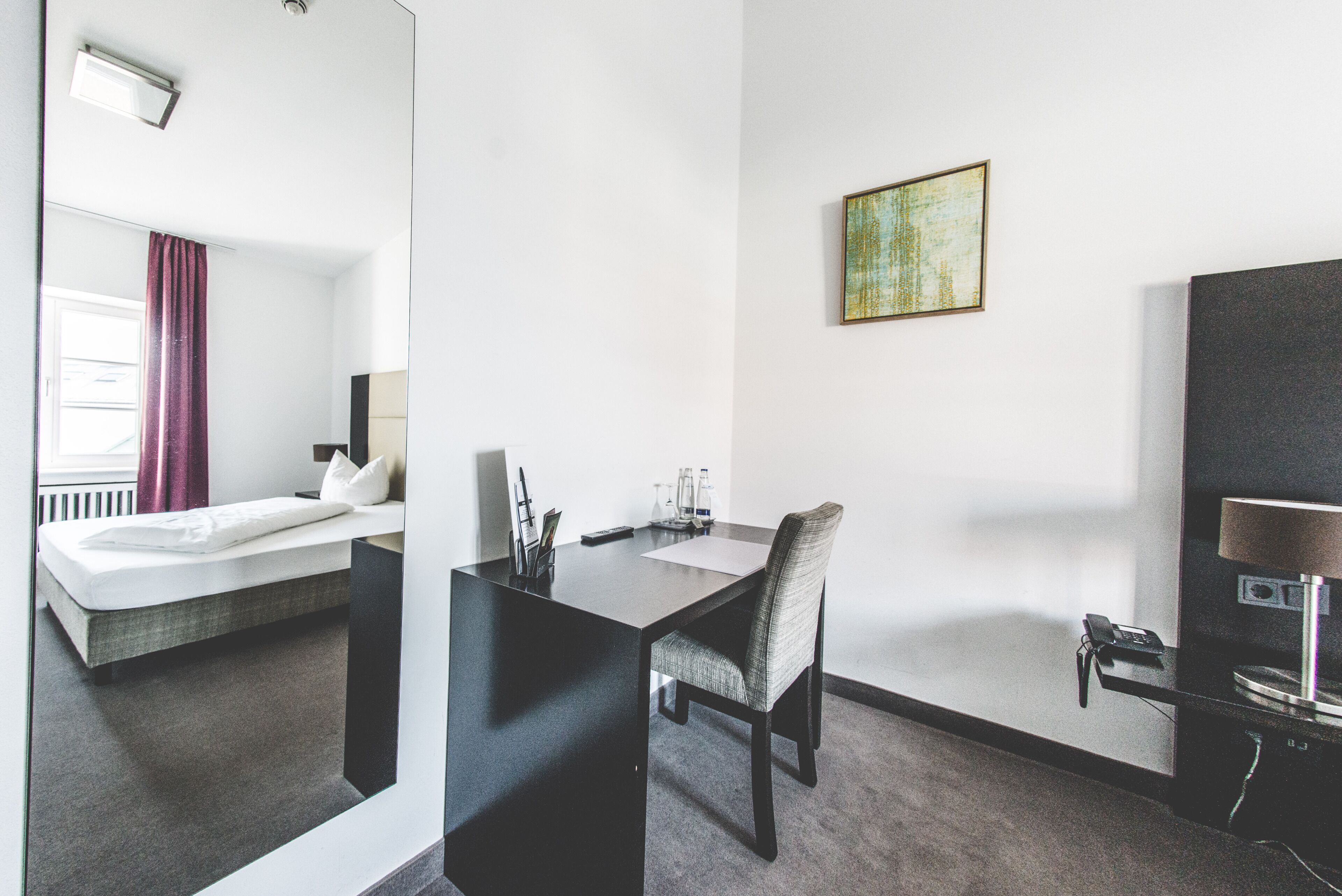 A hotel room with a bed, a desk, a chair, a telephone, and a painting on the wall.