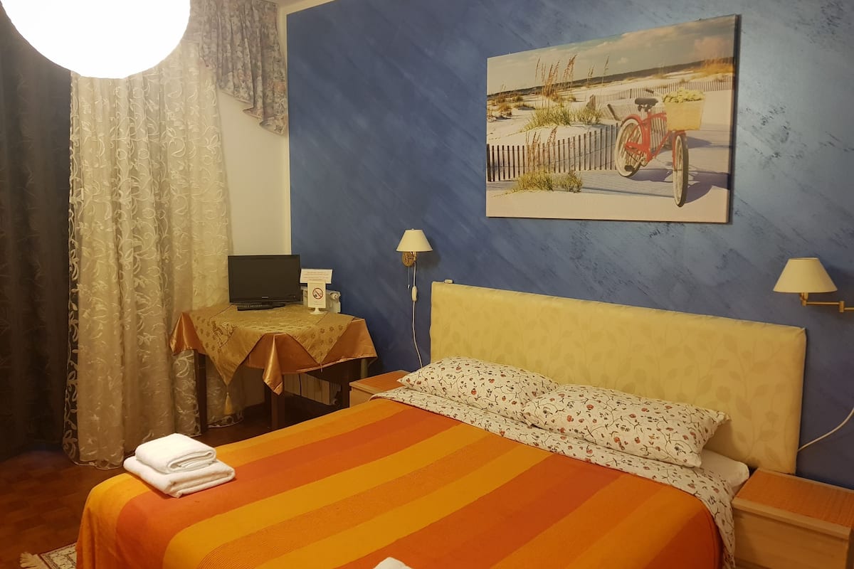 A hotel room with a bed, a desk with a computer, a lamp, and a painting on the wall.