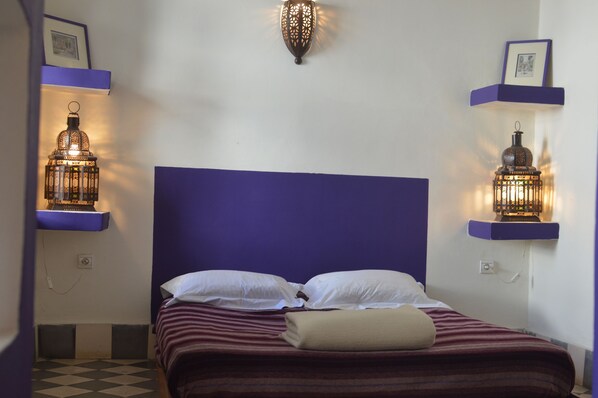 Un lit avec une tête de lit bleue, deux oreillers blancs et une couverture à rayures. Deux lanternes sont fixées aux murs, de part et d’autre du lit.