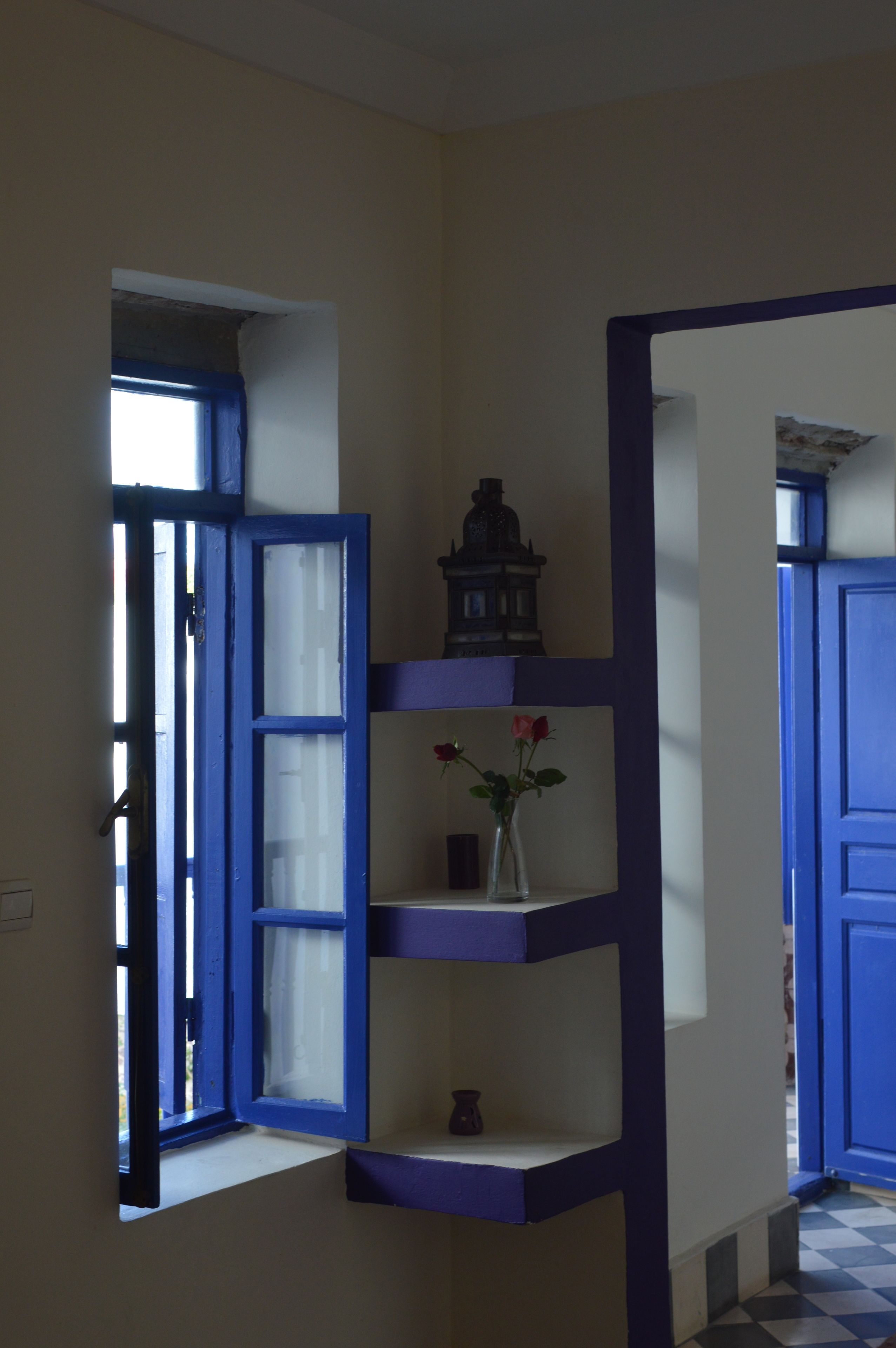 Ein Zimmer mit einem blau gerahmten Fenster, einer kleinen Ablage mit einer Vase und einer Statue sowie einem Schachbrettfußboden.