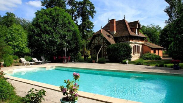Una piscina con sillas de descanso, una casa de estilo Tudor y abundante vegetación.