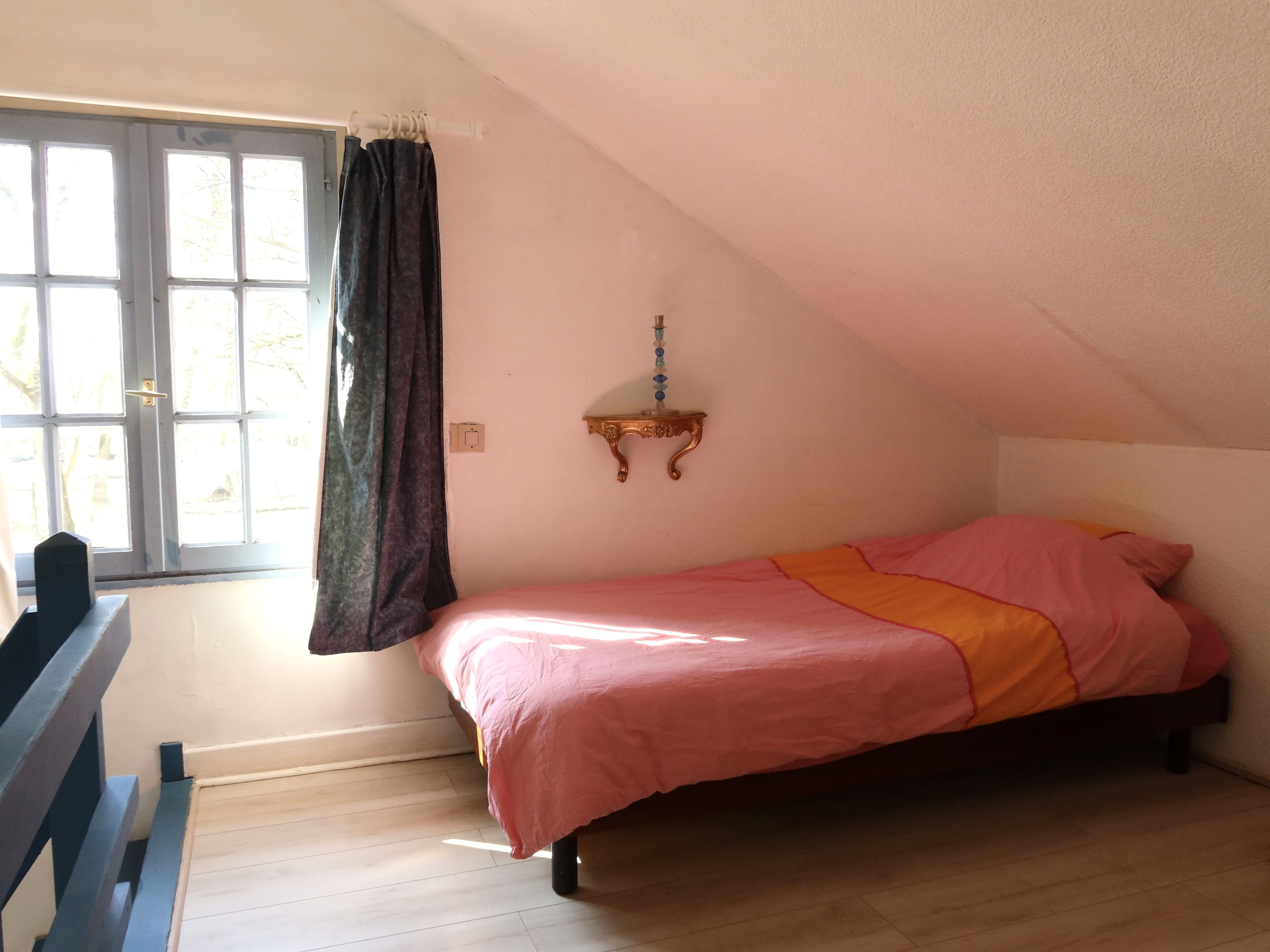 A bedroom with a bed, a window with curtains, a bedside table, and a wall-mounted shelf.