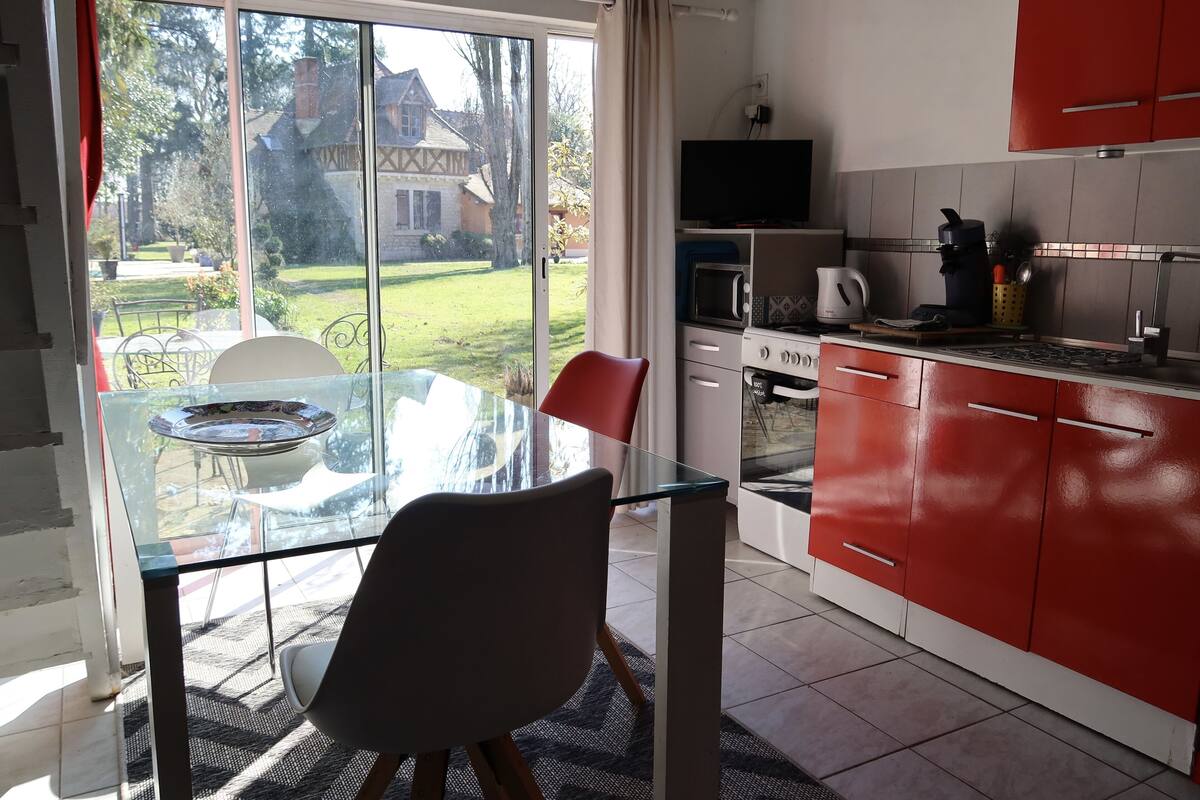 A modern kitchen with red cabinets, a glass dining table, and a view of a garden and house through large windows.