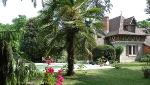 Una casa tradicional con fachada de piedra, un jardín grande con una palmera y un carro de madera lleno de flores.
