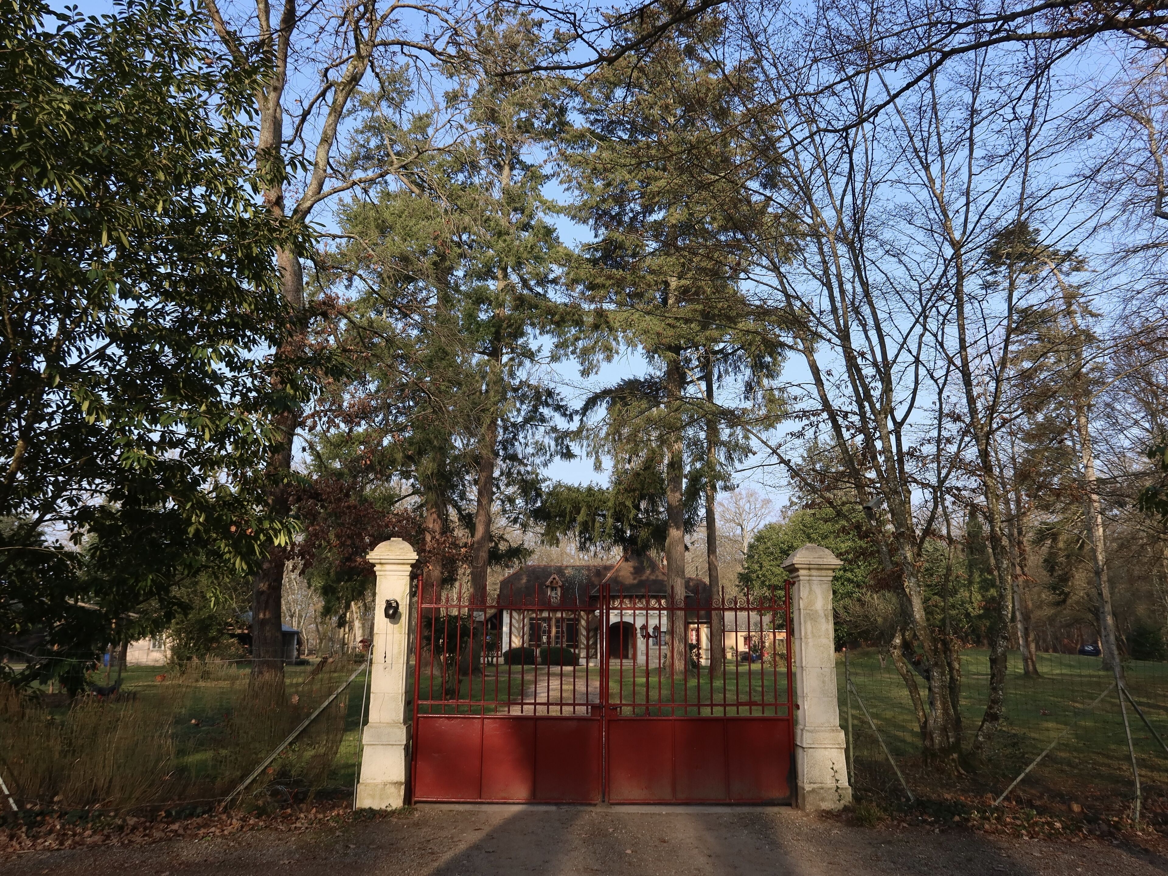 A gated entrance with a stone pillar and a metal gate, surrounded by trees and a grassy area.