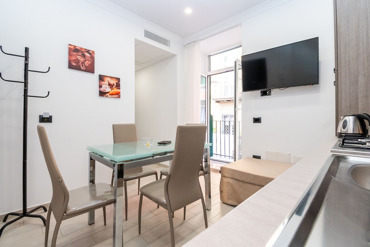 A modern kitchen with a flat-screen TV, a dining area with a glass table and chairs, and a view of the outside balcony.