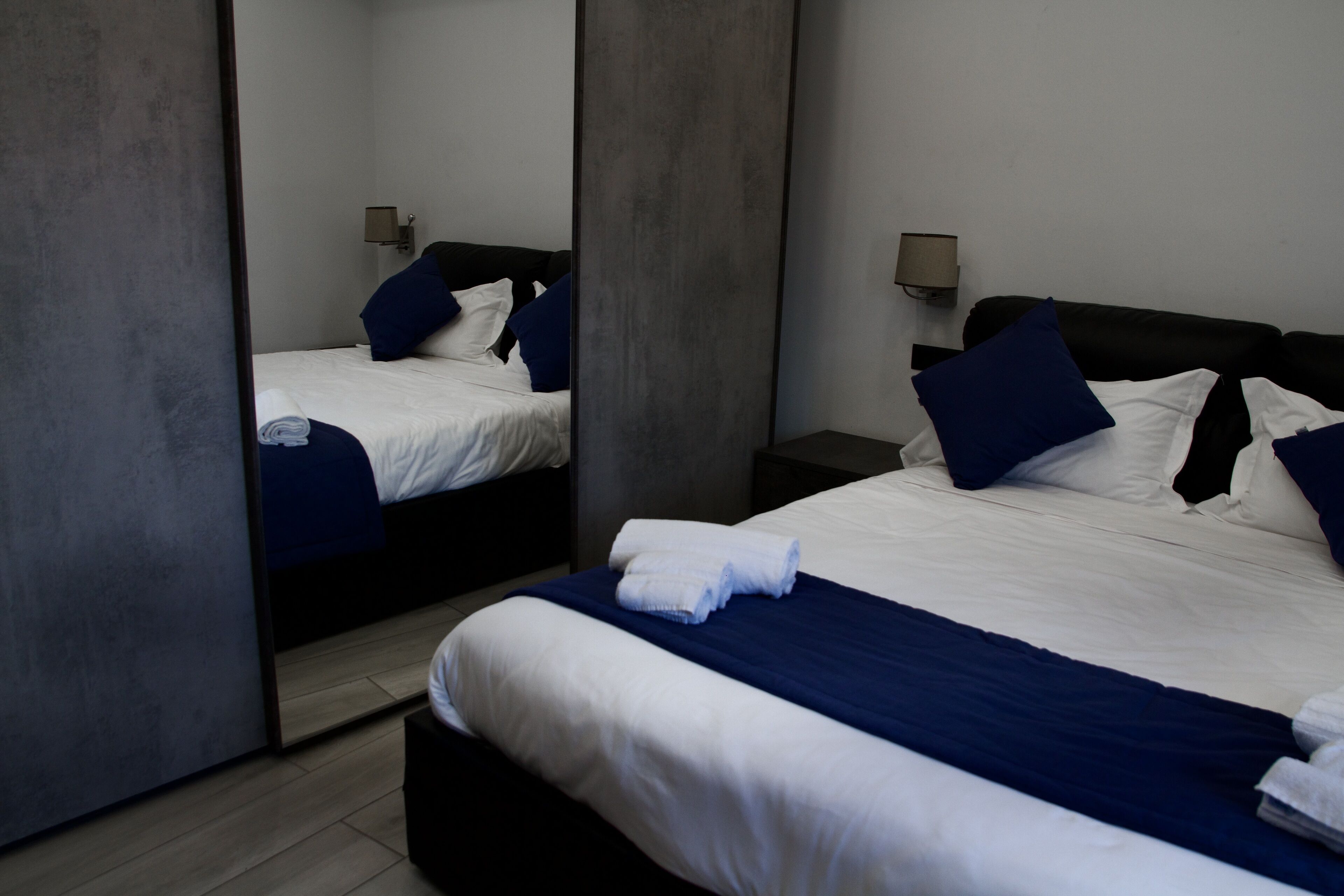 A hotel room with a large mirror, a bed with white and blue bedding, and a nightstand with a lamp.