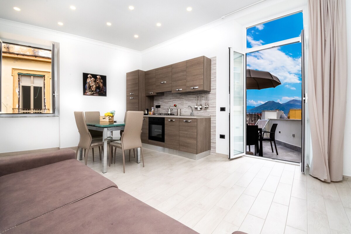 A modern kitchen with wooden cabinets, a dining area with a table and chairs, and a sliding glass door leading to a balcony with a mountain view.