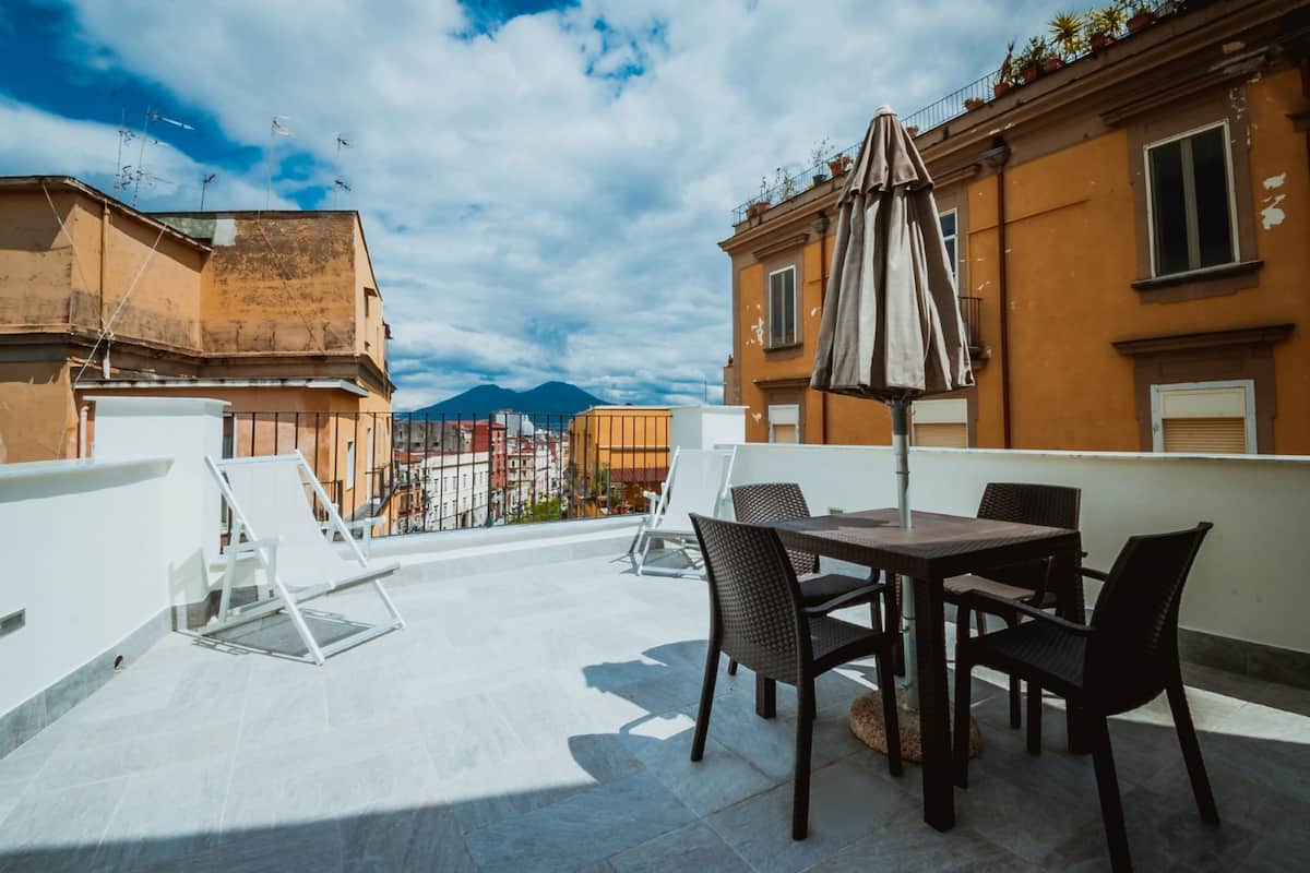 A rooftop terrace with a table and chairs, a sun umbrella, and a view of the city and mountains in the background.