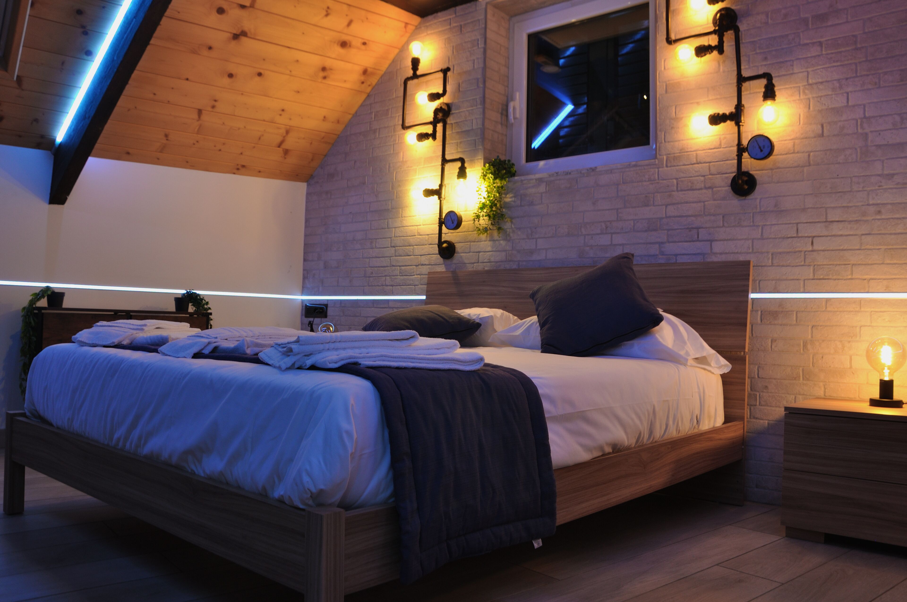 Un dormitorio moderno con techo de madera, pared de ladrillo y cama con ropa blanca y una manta oscura.