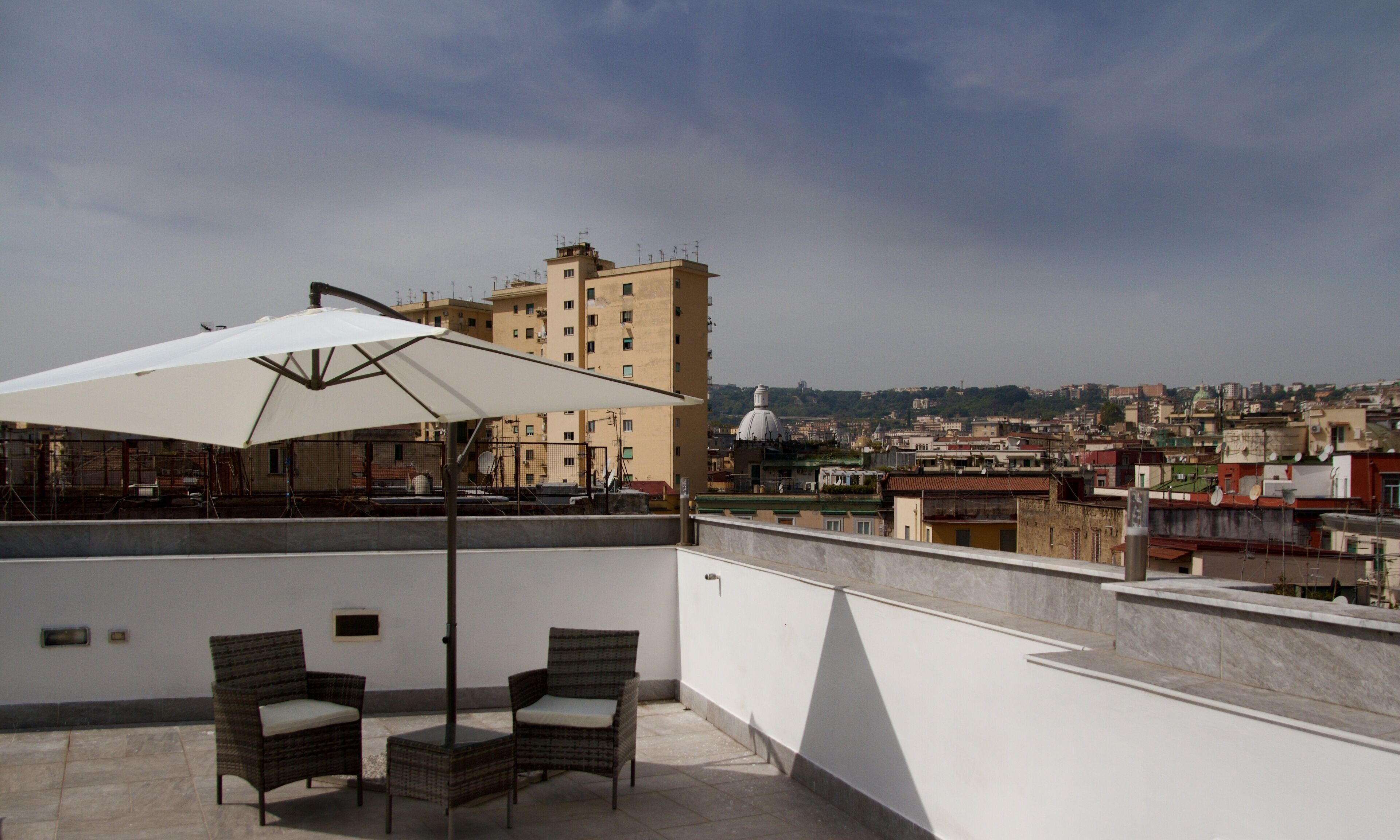 Eine Dachterrasse mit zwei Korbstühlen und einem weißen Sonnenschirm, mit Blick auf eine Stadtlandschaft mit Gebäuden und einer Kuppel in der Ferne.