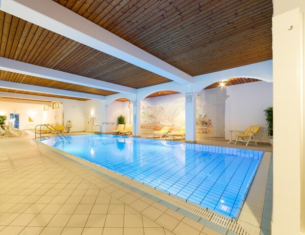 An indoor swimming pool with tiled flooring, lounge chairs, and a wooden ceiling.