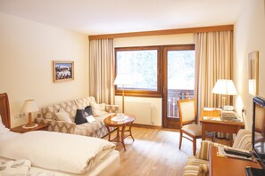 A hotel room with a bed, a sofa, a small table, a chair, a desk with a lamp, and a window with curtains.
