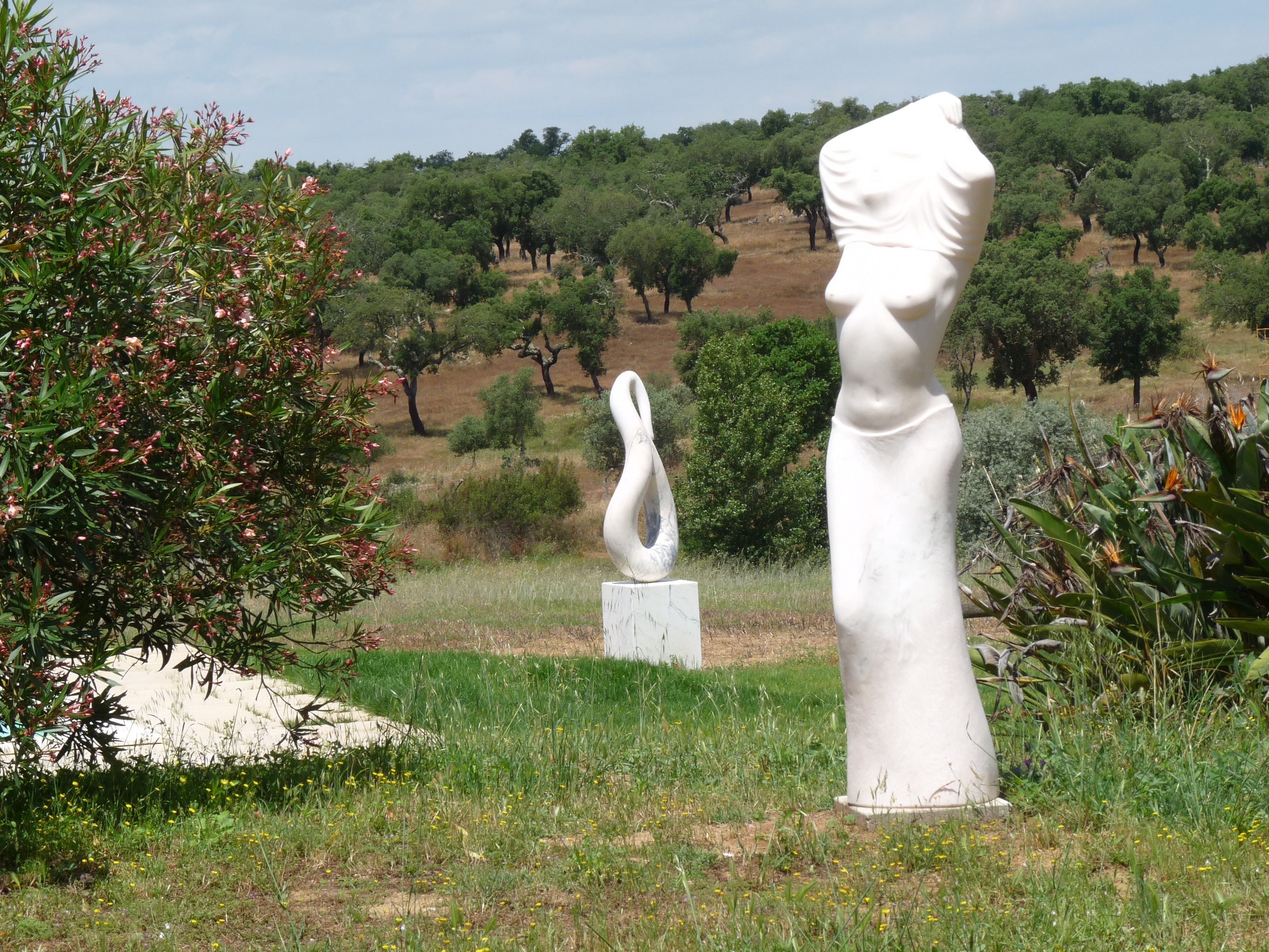 A sculpture garden with two white sculptures, one of a female figure and one of a male figure, surrounded by greenery and trees.