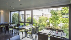 Un restaurante con mesas y sillas dispuestas en el interior, amplias ventanas que ofrecen vistas a zonas verdes y edificios.