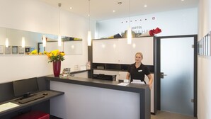 Zona de recepción de un hotel con mostrador de recepción, una computadora, un florero rojo, certificados enmarcados en la pared y un espejo grande.
