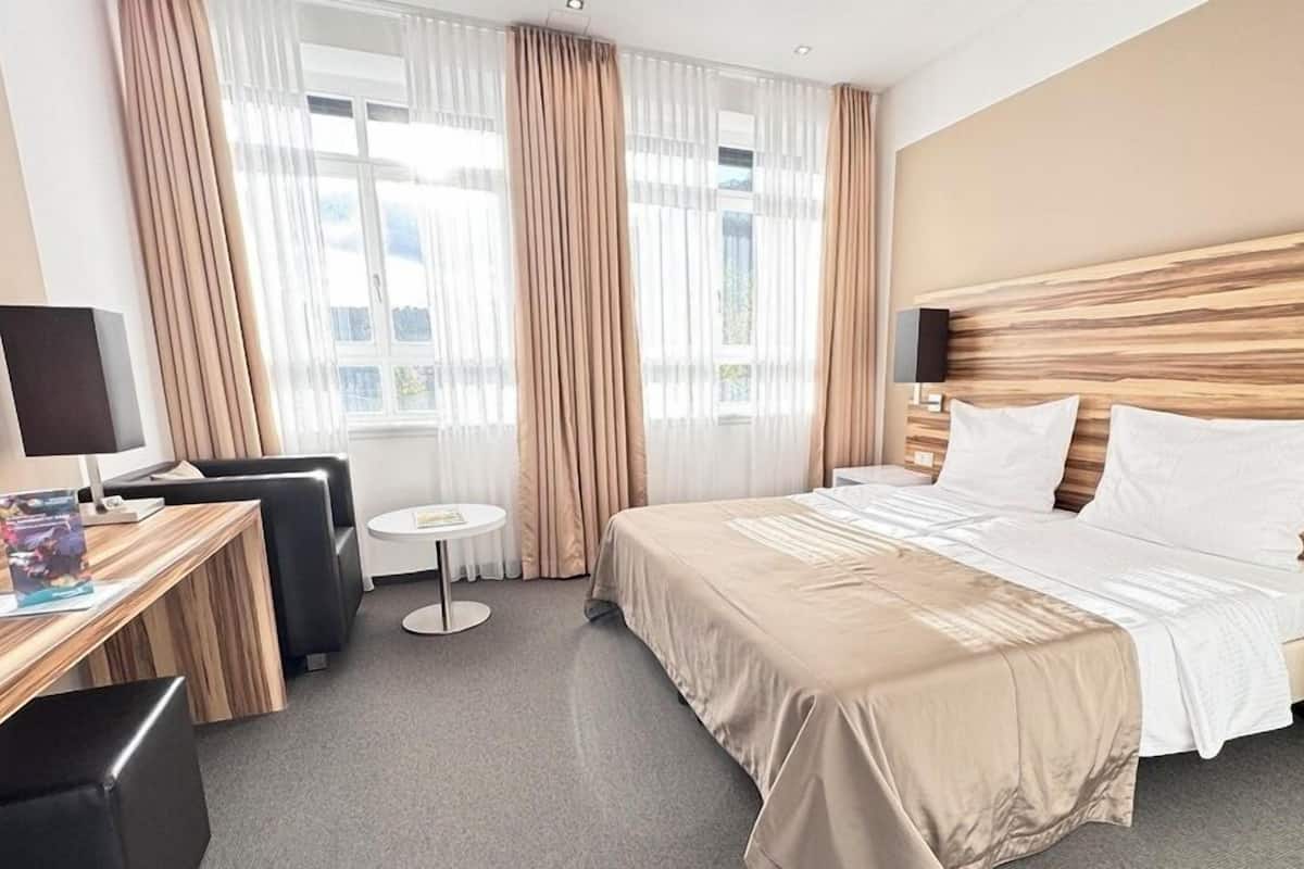 A hotel room with a large bed, a desk with a chair, a small round table, and a window with curtains.