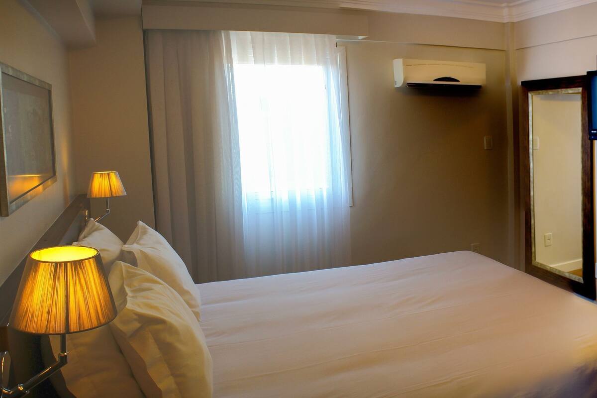 A hotel room with a bed, a lamp, a TV, and a window with curtains.