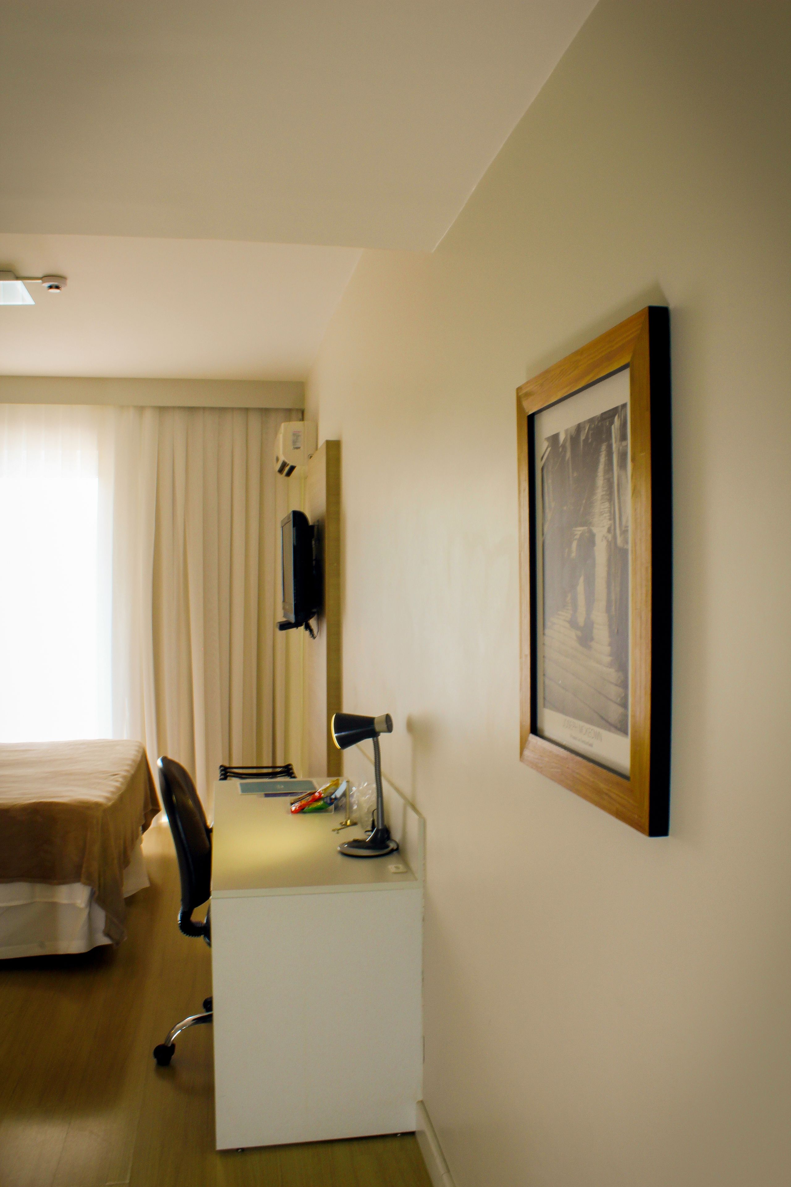 Quarto de hotel com cama, escrivaninha, cadeira e um quadro na parede.