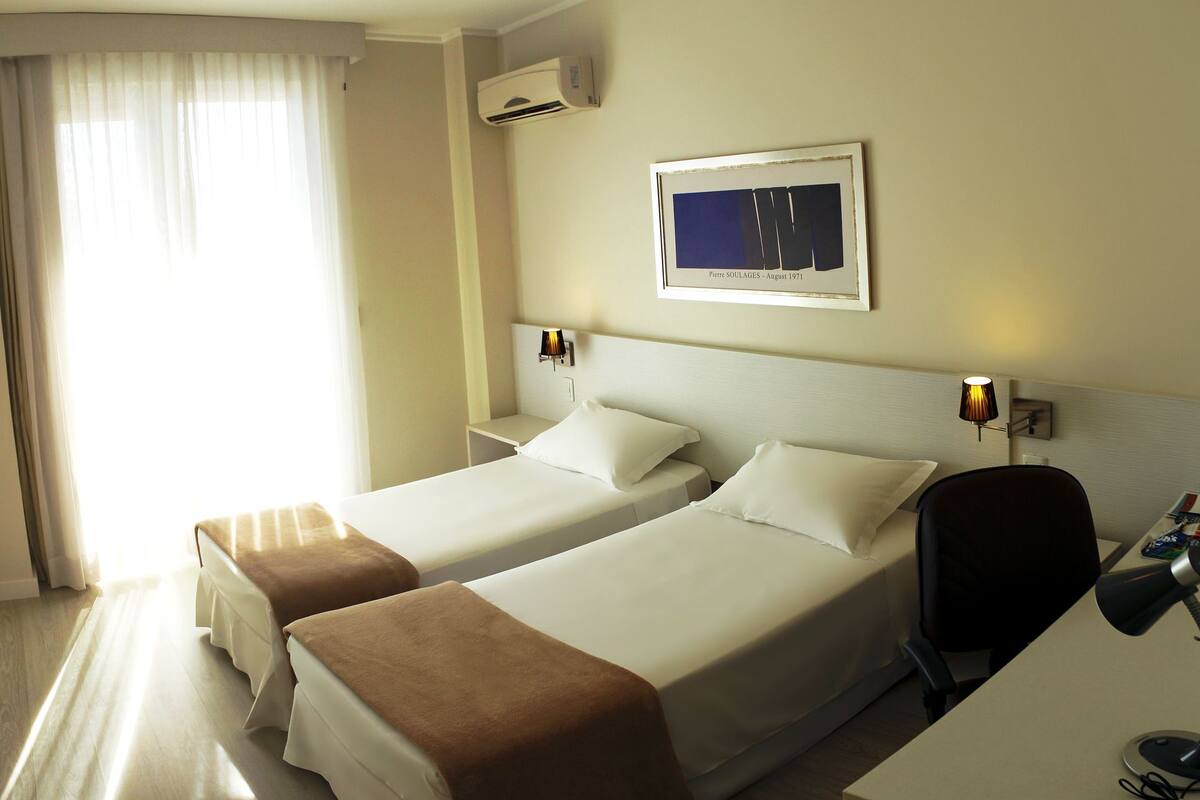 A hotel room with two beds, a desk, and a chair. There is a framed picture on the wall and an air conditioning unit above the bed.