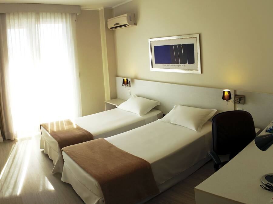 Habitación de hotel con dos camas, un escritorio y una silla. Se observa un cuadro en la pared y un equipo de aire acondicionado sobre la cama.