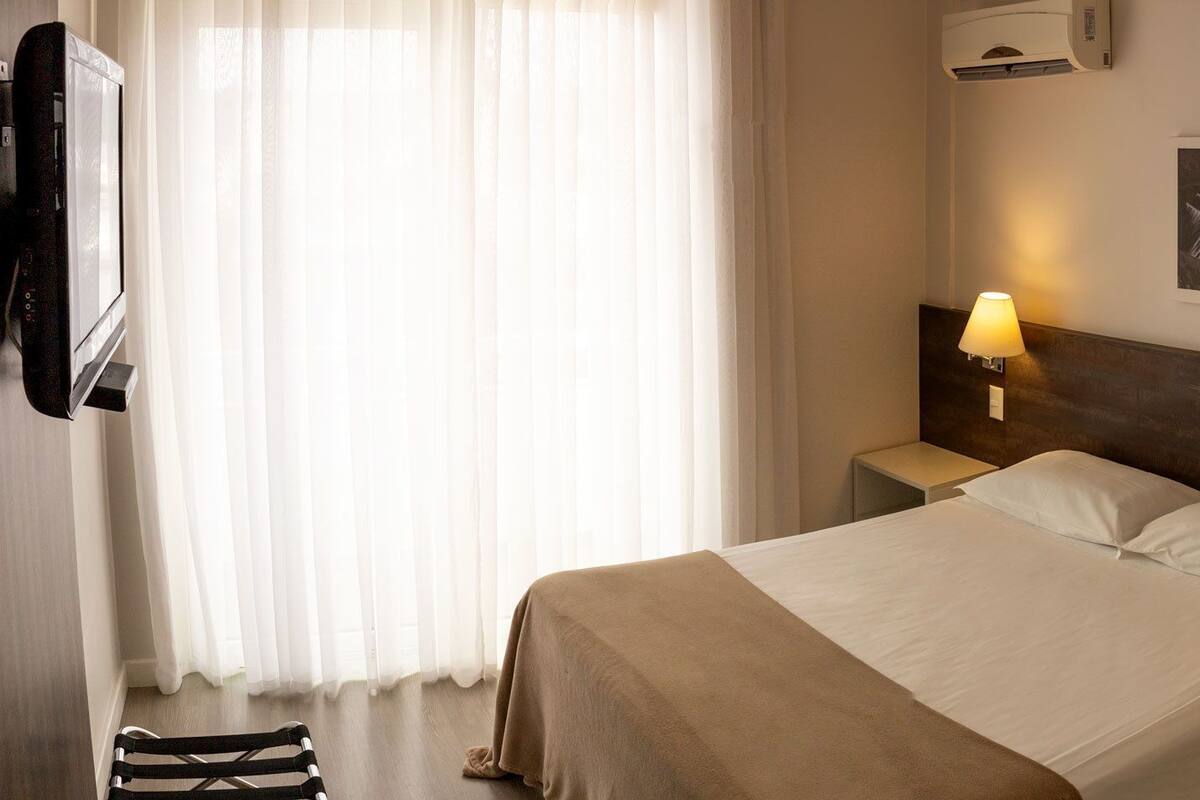 A hotel room with a bed, two bedside lamps, a telephone, a wall-mounted air conditioner, and a framed picture.