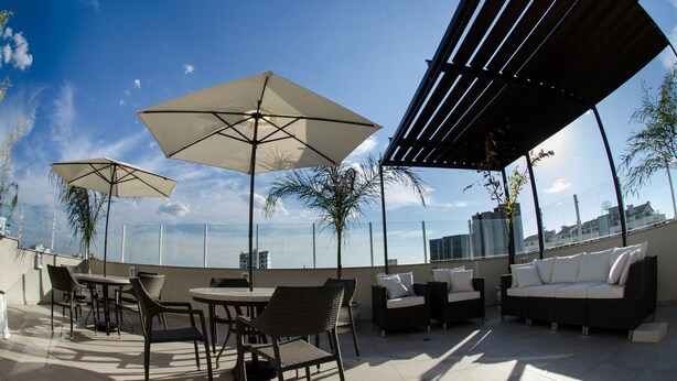 Un patio al aire libre con mesas, sillas y un sofá bajo una pérgola, con vista a un paisaje urbano.