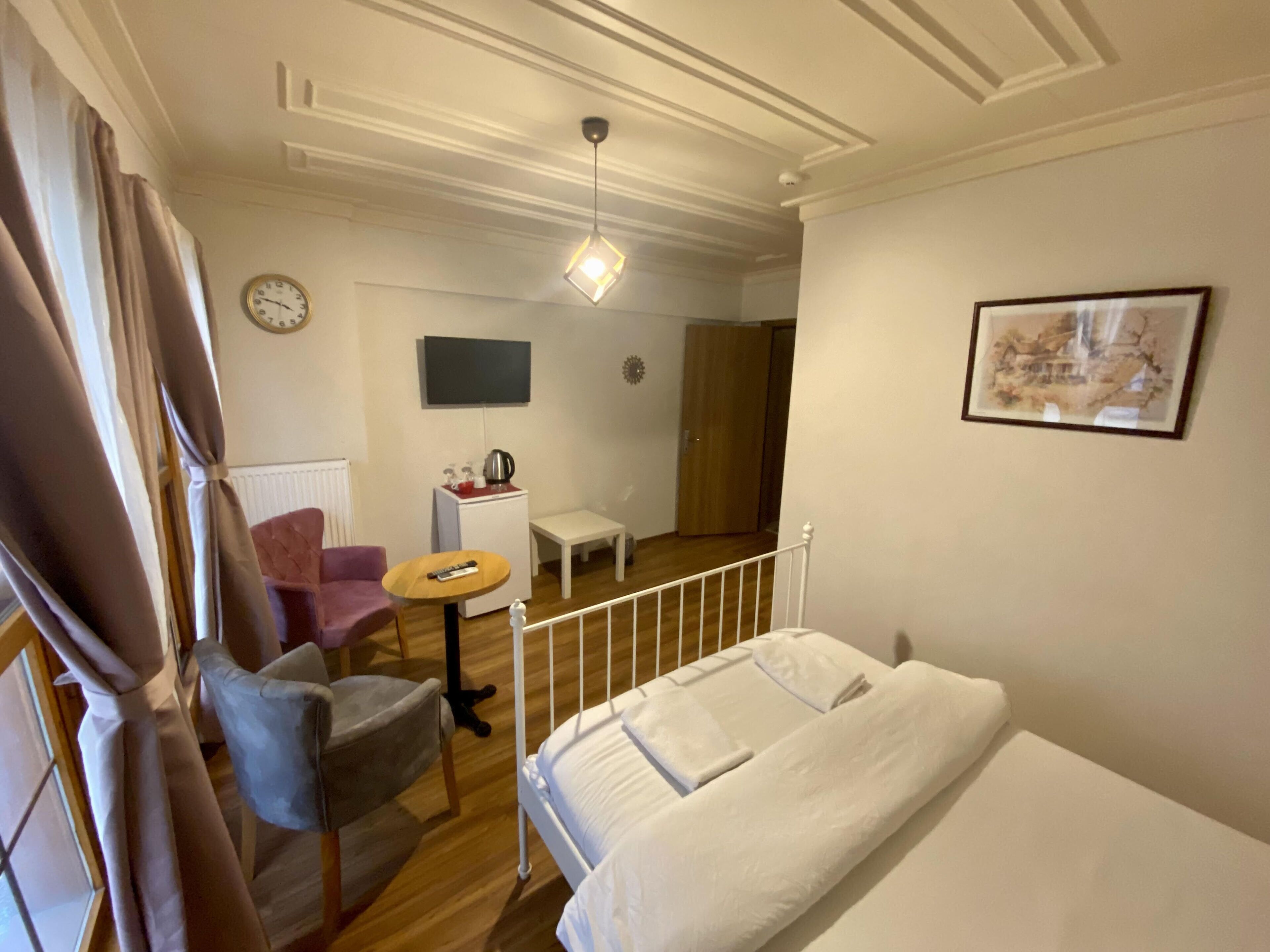 A hotel room with a bed, a chair, a small table, a TV, a clock, and a door leading to another room.
