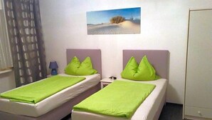 A room with two beds, each with a green pillow and a white bedspread. There is a nightstand with a lamp between the beds. A painting of a desert landscape hangs on the wall above the beds.