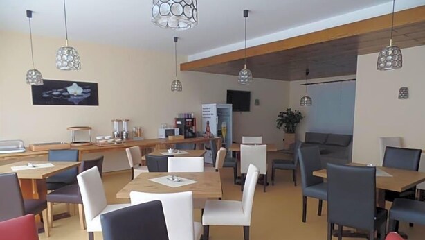 A modern dining area with multiple tables and chairs, pendant lighting, and a flat-screen TV mounted on the wall.