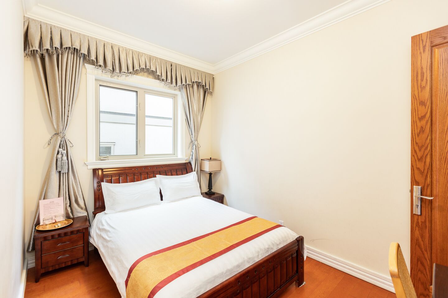 A neatly made bed with a wooden headboard, a bedside table with a lamp, and a window with curtains.