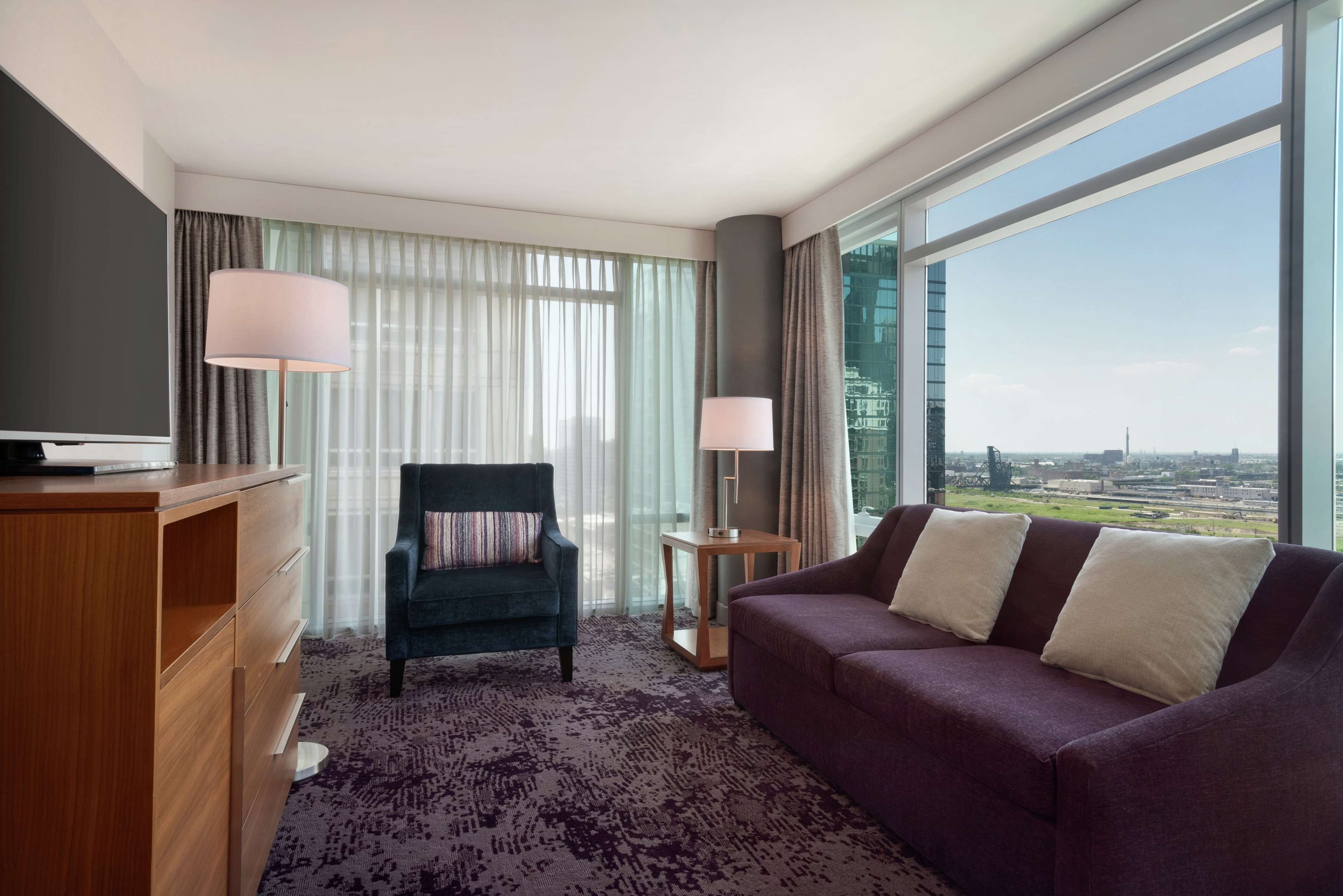 Una habitación de hotel moderna con un ventanal grande, un sofá púrpura, un escritorio de madera y una silla.