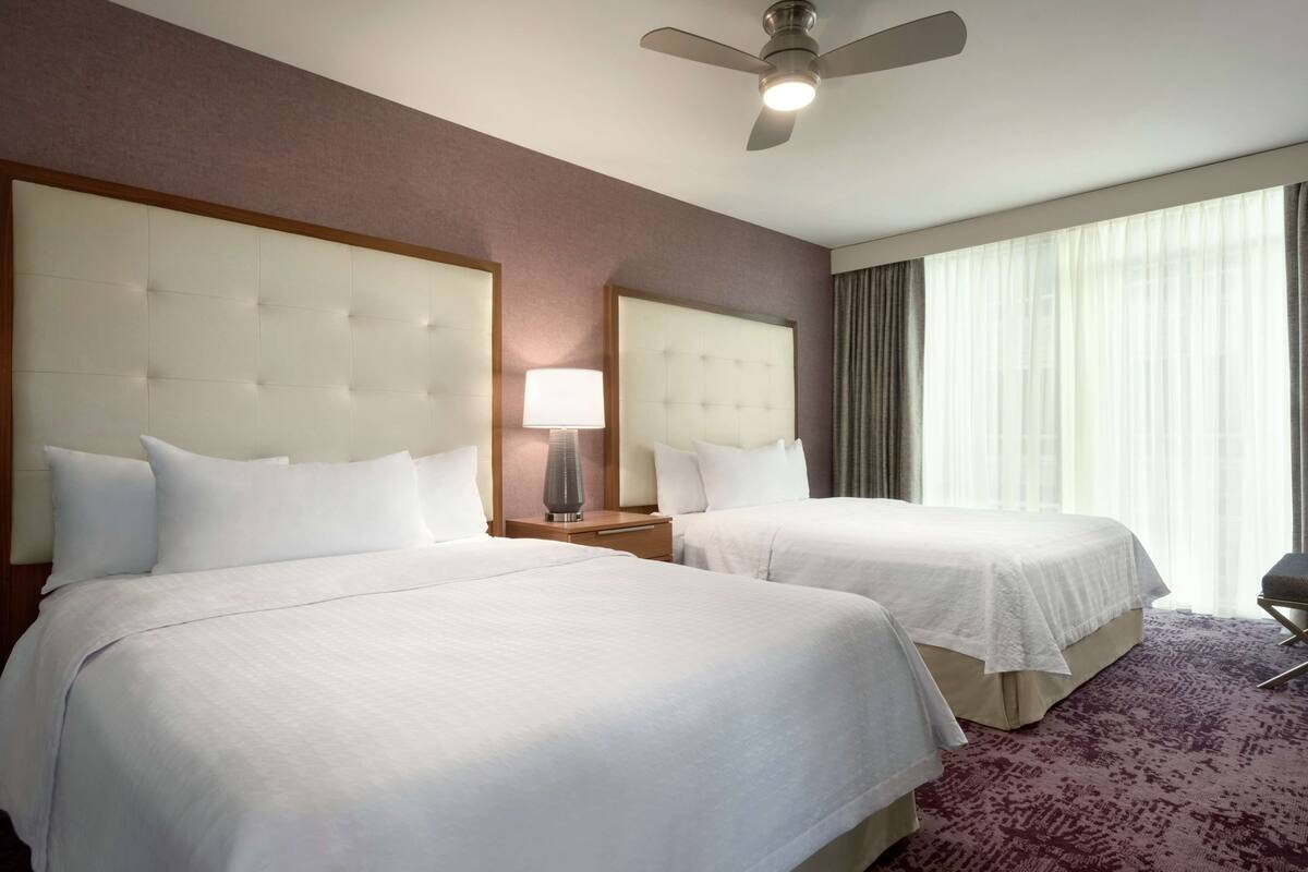 A hotel room with two beds, a ceiling fan, a nightstand with a lamp, and a large window with curtains.