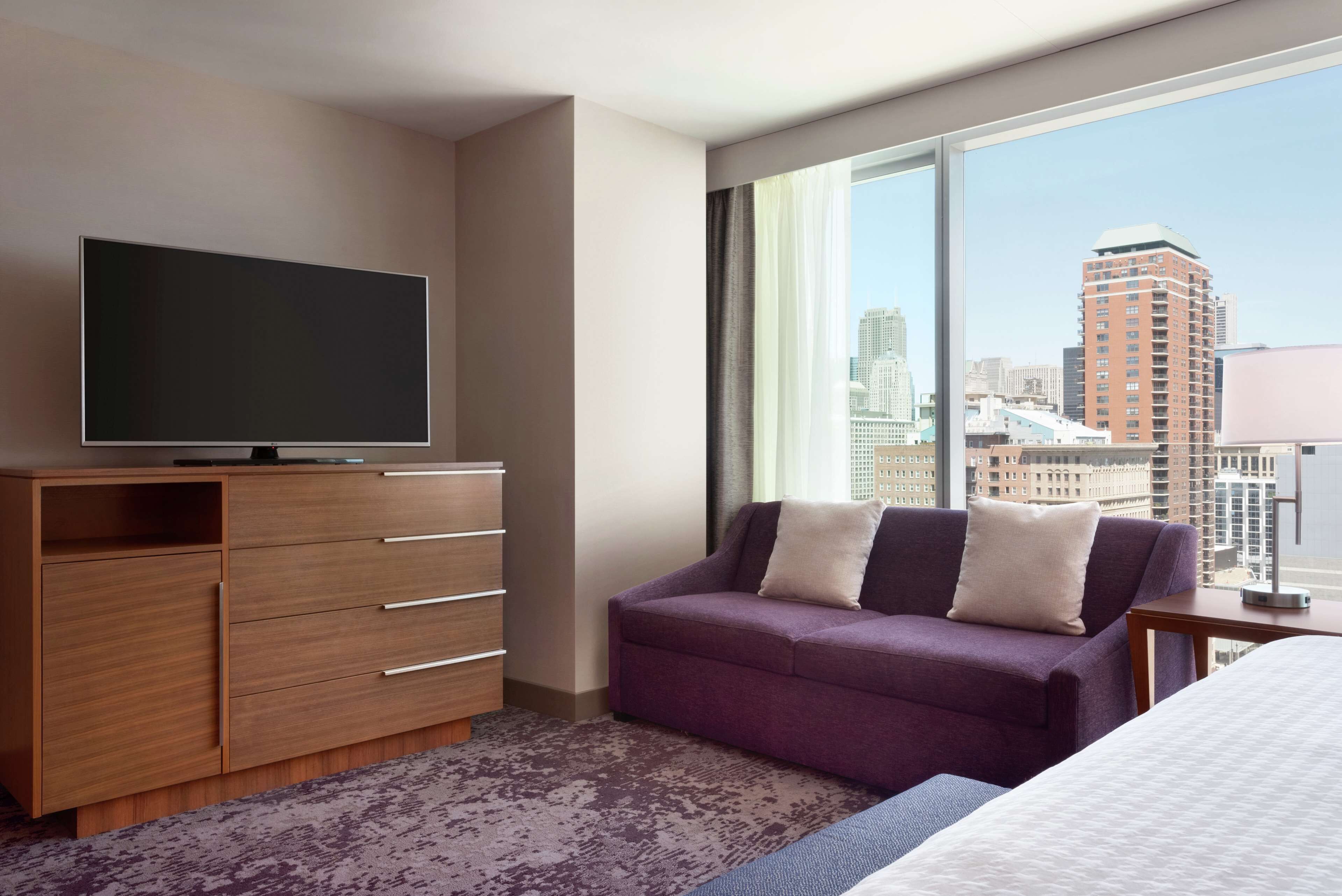 A hotel room with a large window, a flat-screen TV, a purple sofa, and a bed.