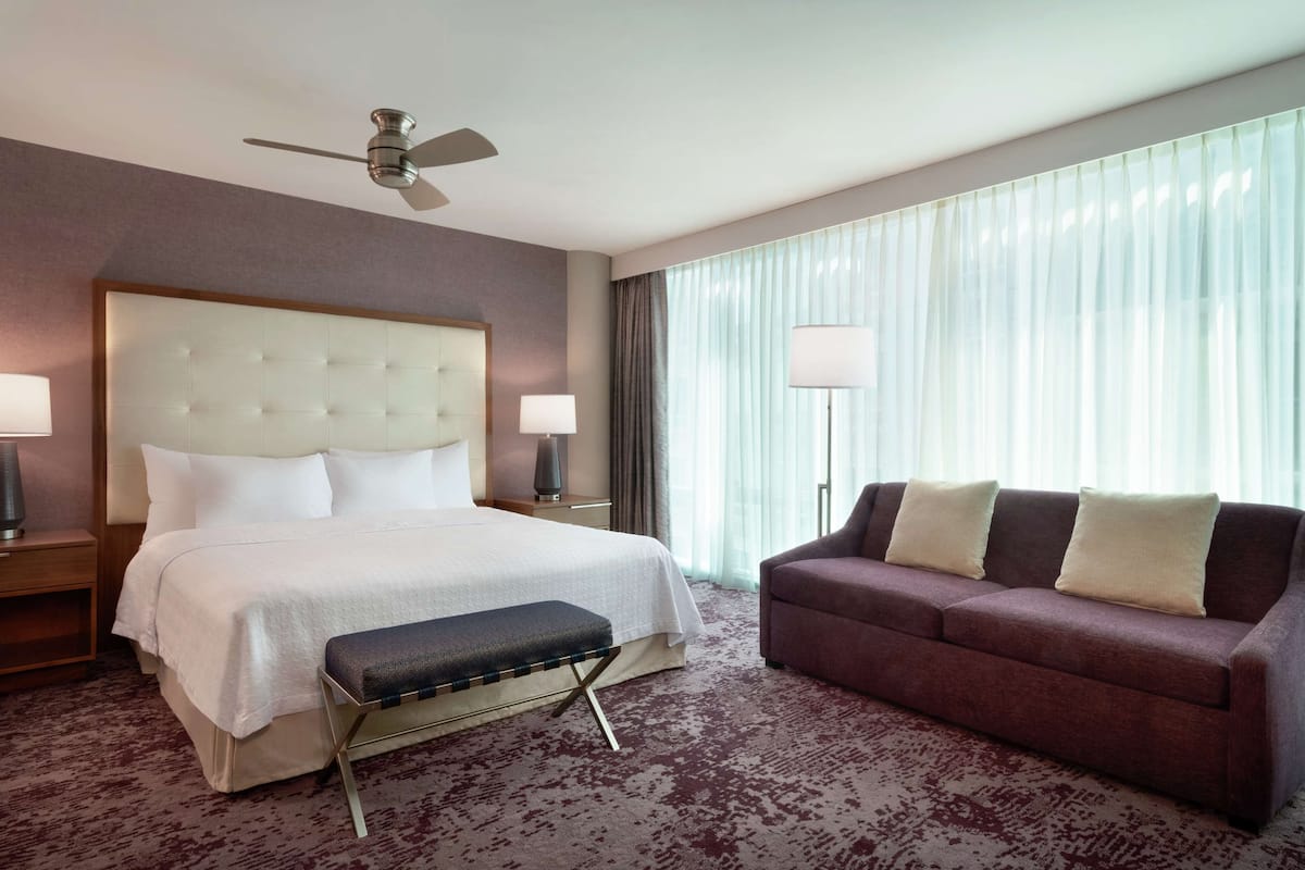 A hotel room with a large bed, a sofa, a ceiling fan, and a large window with curtains.
