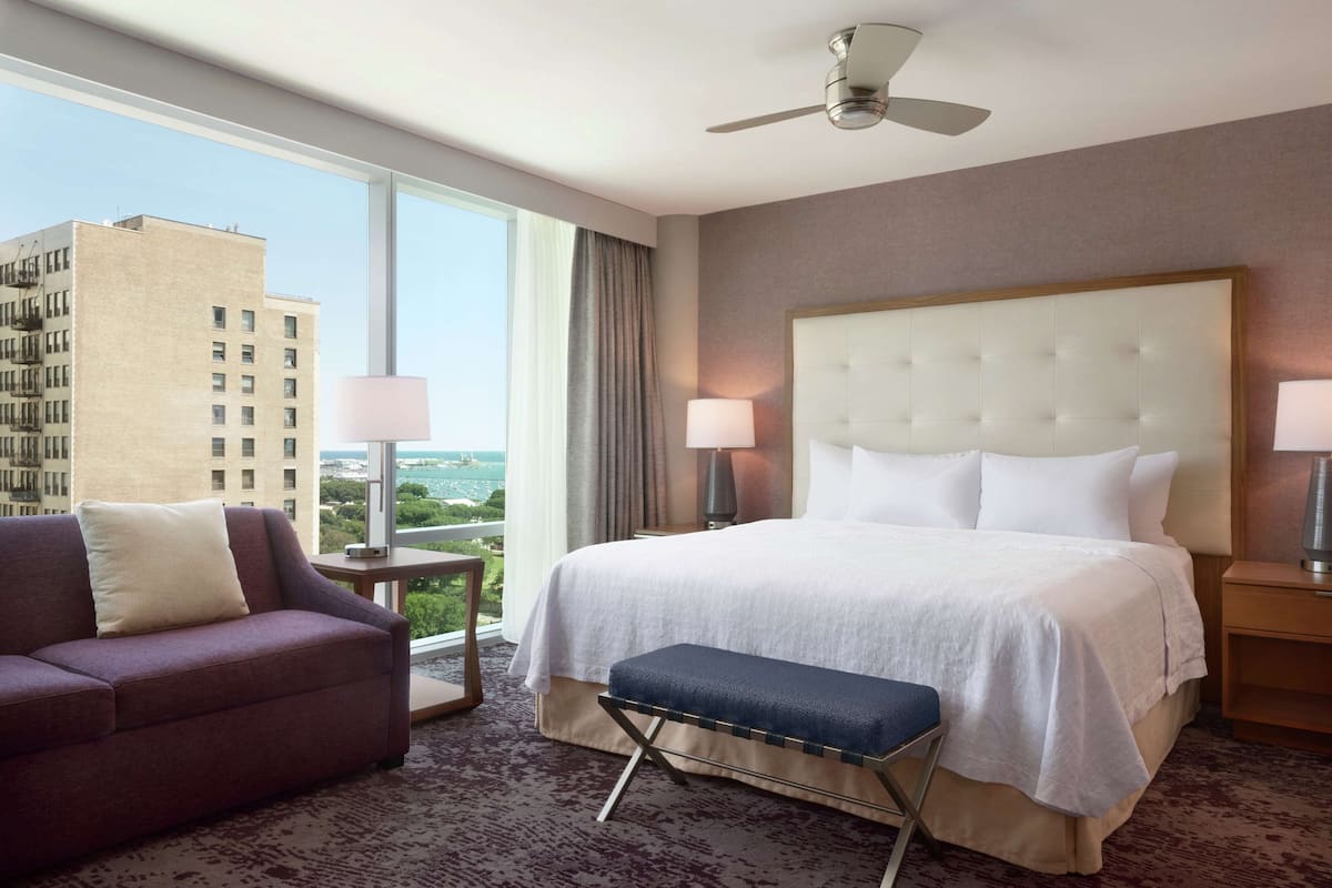 A hotel room with a large bed, a sofa, a bench, a ceiling fan, and a view of a cityscape through large windows.