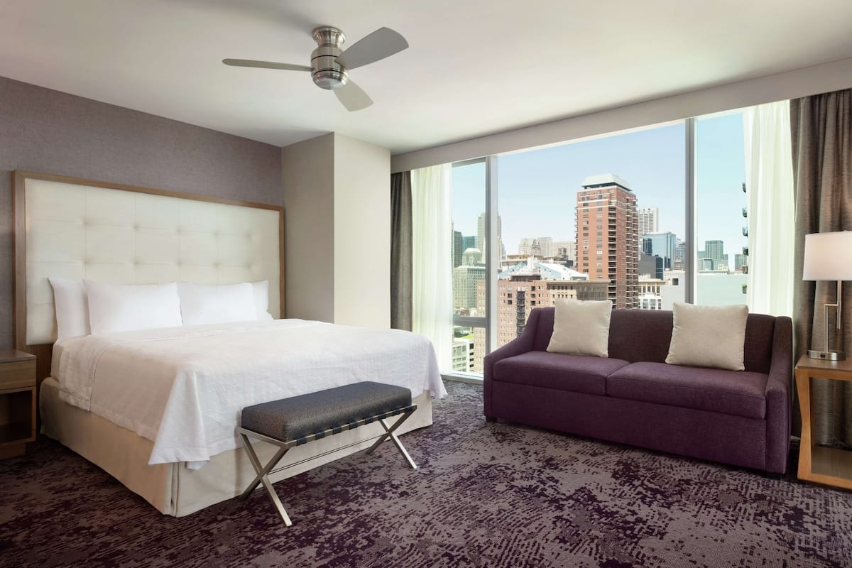 A hotel room with a large bed, a bench, a purple sofa, and a city view through large windows.