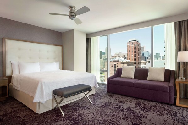 Habitación de hotel con una cama grande, un banco, un sofá púrpura y vistas a la ciudad a través de amplias ventanas.